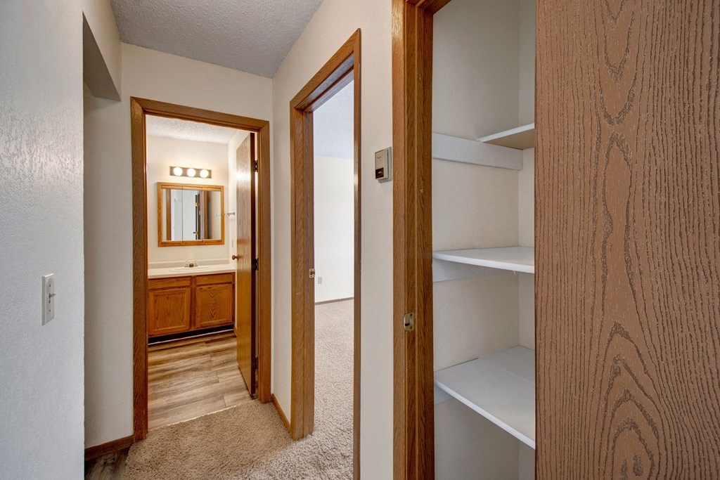 Narrow hallway here at Windscape Apartment Homes with neutral tones, featuring a door to a well-lit bathroom with a large mirror and wooden cabinet. Open closet with shelves on the right. Cozy atmosphere.