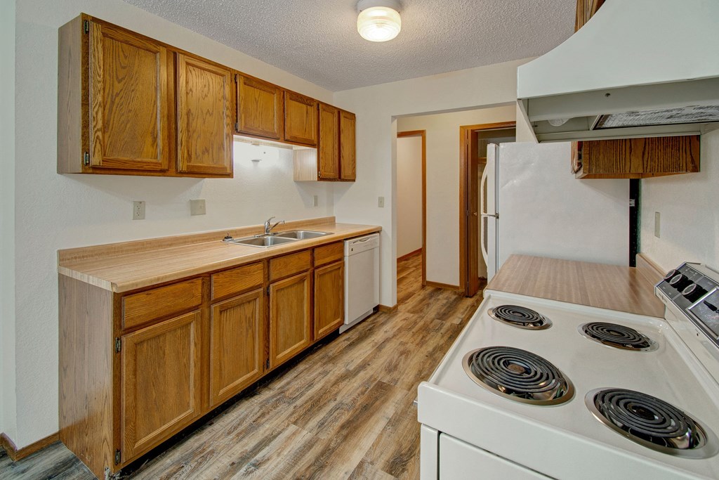 Narrow kitchen here at Windscape Apartment Homes with wood cabinets, white appliances, and a dual sink. Bright ceiling light, natural wood flooring, and minimalist modern tone.