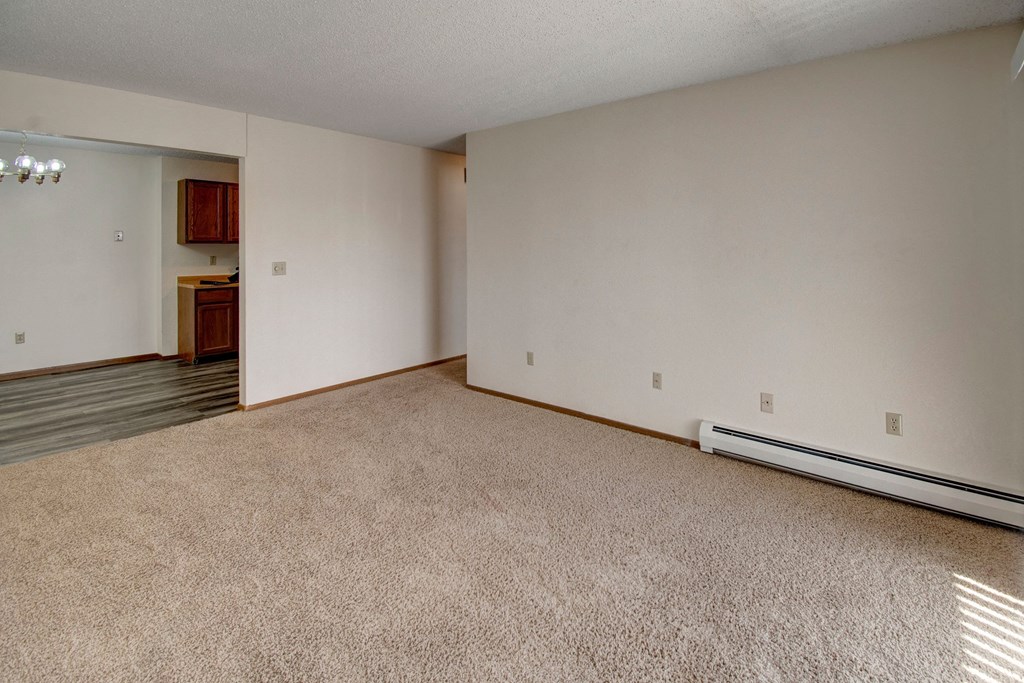 An empty living room here at Windscape Apartment Homes with beige carpet, cream walls, and a visible baseboard heater. An open doorway leads to a kitchen with wooden cabinets and laminate flooring.