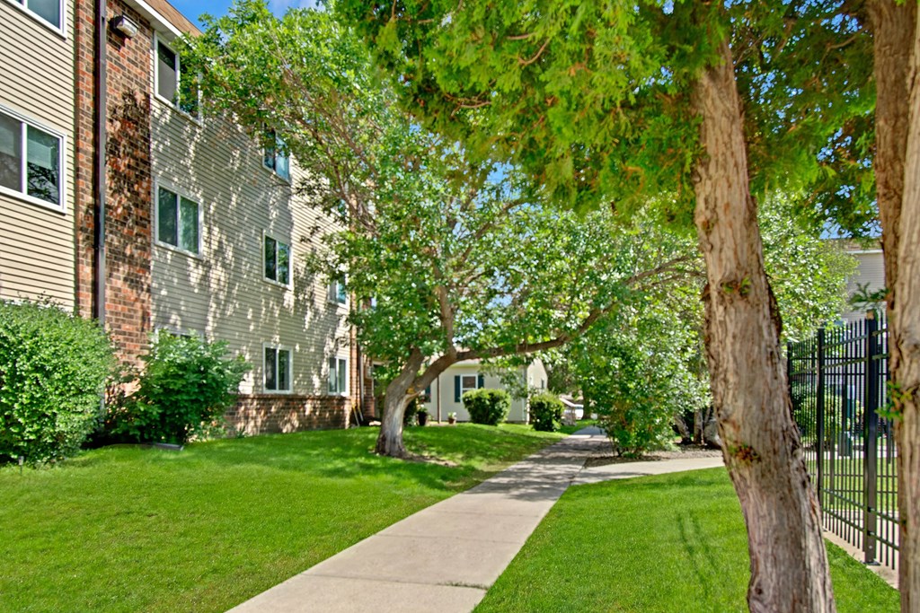 A sidewalk lined with lush green grass and trees leads past an apartment building with beige siding and brick accents here at Windscape Apartment Homes. The scene is tranquil and sunny.
