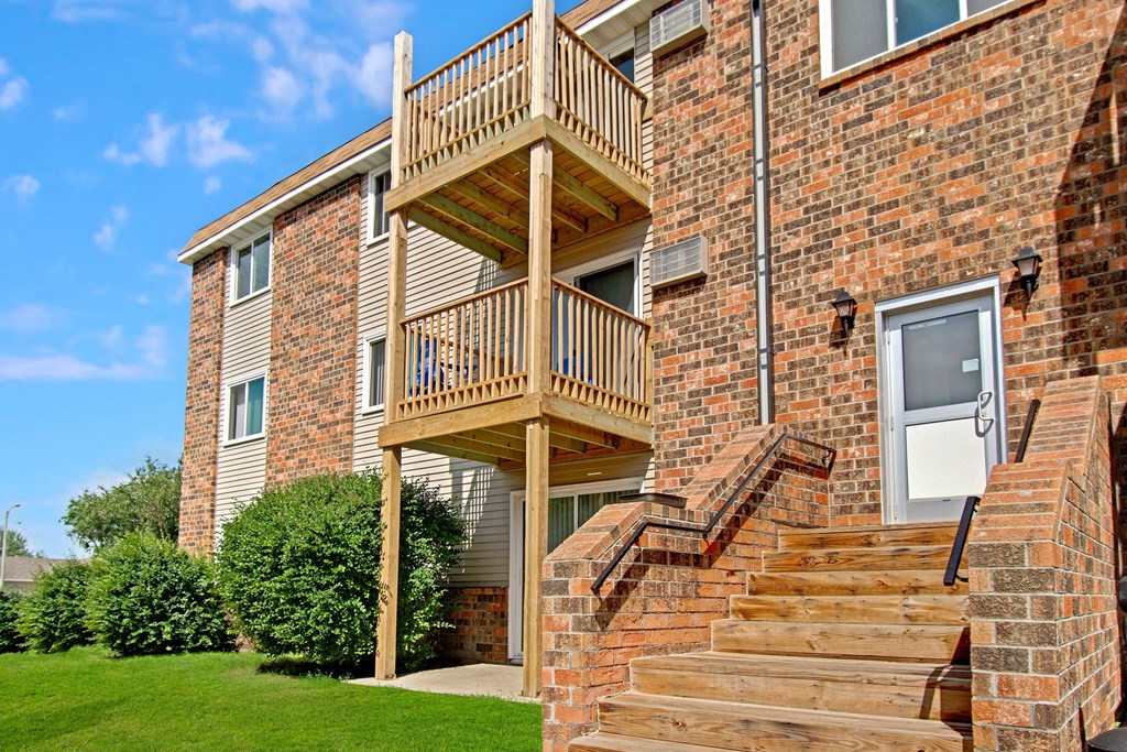 Apartment exterior here at Windscape Apartment Homes with red brick walls and two wooden balconies. Wooden stairs lead to a white door. Green bushes line the building, and the sky is blue.
