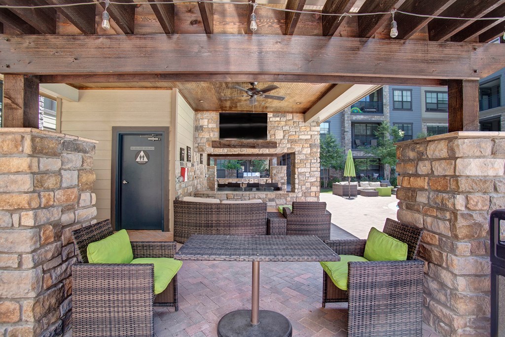 Covered patio area here at the Enclave at Brookside Apartments with wicker furniture and lime green cushions. Stone walls and wooden beams complement a TV above a fireplace. Relaxed, inviting atmosphere.