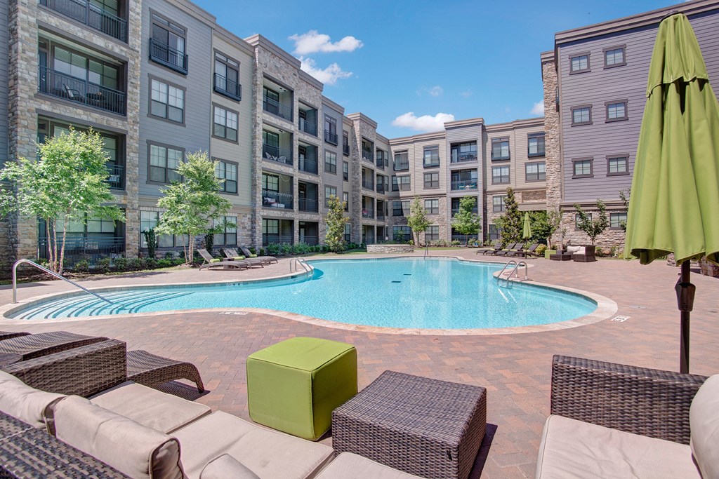 Spacious swimming pool surrounded by the Enclave at Brookside's modern apartment complex under a clear blue sky. Wicker furniture with green cushions adds a cozy touch.