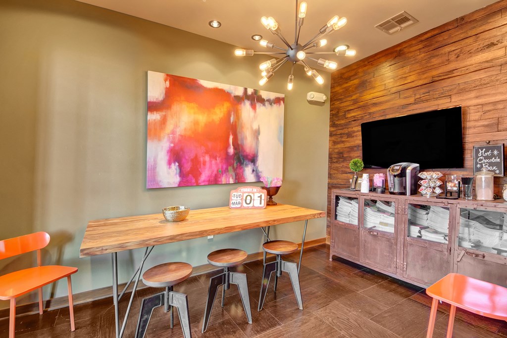 Modern coffee room here at the Enclave at Brookside Apartments with a wooden table, three stools, and orange chairs. Abstract red-and-white art adorns the wall. A cabinet holds coffee supplies. Warm lighting and a cozy atmosphere.