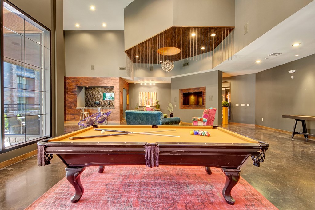 Spacious lounge here at the Enclave at Brookside Apartments with a central pool table on a red rug. Modern decor, high ceilings, large windows, comfortable seating, and a cozy atmosphere.