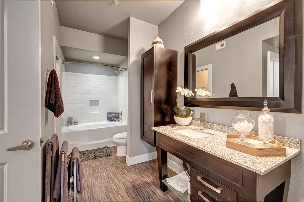 Modern bathroom here at the Enclave at Brookside Apartments with dark wood accents, featuring a granite-topped vanity with a large mirror. A white bathtub has a gray mat, and the floor is wooden. Elegant and clean design.