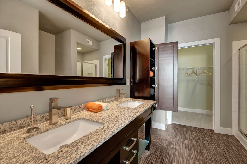 Modern bathroom here at the Enclave at Brookside Apartments with a granite double vanity, sleek faucets, and a large mirror. An open wooden cabinet reveals neatly folded towels. A walk-in closet is visible through an open door.