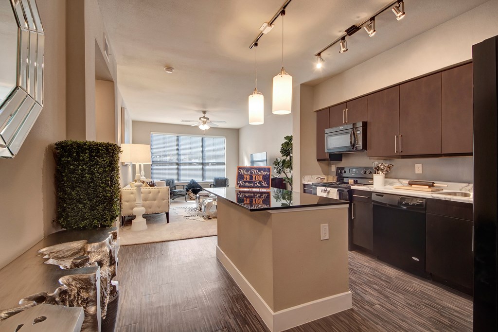 Modern open-concept kitchen and living area here at the Enclave at Brookside Apartments with dark wood cabinets, stainless steel appliances, pendant lighting, and a cozy seating nook by large windows.