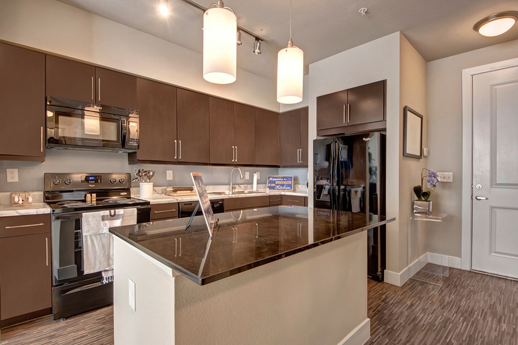 Modern kitchen here at the Enclave at Brookside Apartments with dark wood cabinets, black appliances, and granite countertops. Two pendant lights hang above a central island. Light and minimalist tone.