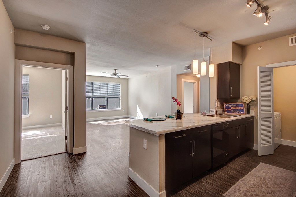 Spacious modern apartment here at the Enclave at Brookside Apartments with dark wood floors, a kitchen island, pendant lights, and a bright living area. Open door reveals an adjacent room.