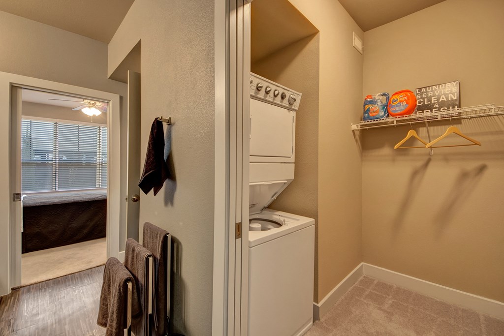 Stacked washer and dryer in a compact laundry area here at the Enclave at Brookside Apartments, beige walls, shelf with detergent, and hangers. Open door reveals bedroom with large window. Cozy ambiance.
