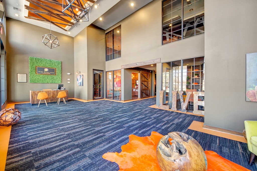 Spacious lobby with high ceilings here at the Enclave at Brookside Apartments, modern geometric lighting, and a mix of blue carpet and orange accents. Wooden decor pieces and casual seating create a welcoming atmosphere.