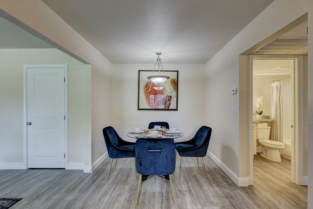 Indigo Apartments Dining Room Cozy dining area here at Indigo Apartment Homes with a round table set for four, surrounded by navy velvet chairs. A modern abstract painting hangs above, and soft lighting creates a warm ambiance.
