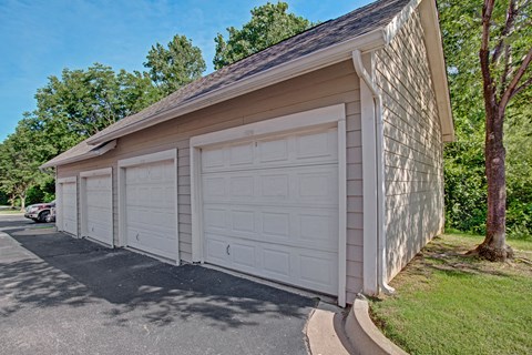 A row of three closed, white garage doors on a beige building under clear skies, surrounded by lush green trees, here at Memorial Creek Apartment Homes. The mood is calm and orderly.