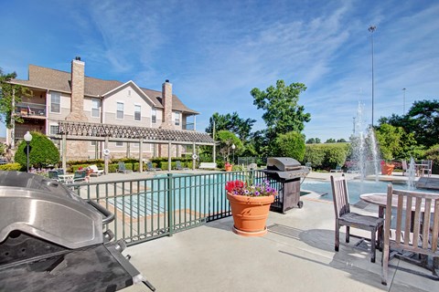 Sunny pool area here at Memorial Creek Apartment Homes with a brick house, barbecue grills, wooden tables, and chairs. Fountains spray water near potted flowers, set against a clear blue sky.