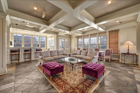 Elegant resident lounge here at Memorial Creek Apartment Homes with coffered ceiling, beige sofas, purple tufted ottomans, glass coffee table, warm lighting, and large windows; cozy and inviting.