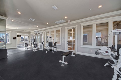 Bright gym interior here at Memorial Creek Apartment Homes with modern fitness equipment, including weight machines and a benched area. Large windows allow natural light, creating an inviting atmosphere.