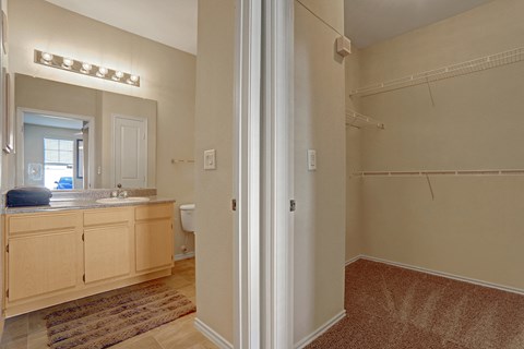 Beige bathroom and empty closet here at Memorial Creek Apartment Homes. The bathroom features a vanity with a large mirror and bright lights, while the closet has carpet and wire shelves.