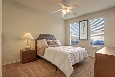 Cozy bedroom here at Memorial Creek Apartment Homes with a neatly made bed, beige and brown linens, nightstand with lamp, and floral artwork. Two windows provide natural light.