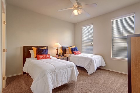 Bright, cozy twin bedroom here at Memorial Creek Apartment Homes with neutral walls and carpet. Two beds have white bedding, colorful pillows, and a shared lamp-lit nightstand, conveying warmth.