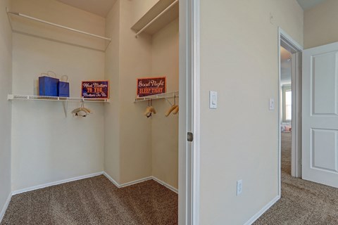 Walk-in closet here at Memorial Creek Apartment Homes with beige walls and brown carpet, containing blue gift bags, motivational signs, and wooden hangers. Open door leads to a sunlit room.