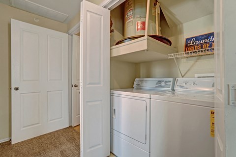 A compact laundry room here at Memorial Creek Apartment Homes with a washer and dryer, partially hidden by open white doors. Above, there’s a water heater and a sign reading "Laundry Loads of Fun."