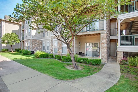 Memorial Creek Apartment Homes with stone and beige siding, featuring lush green lawns, a shady tree, and a paved walkway. Bright, welcoming atmosphere.