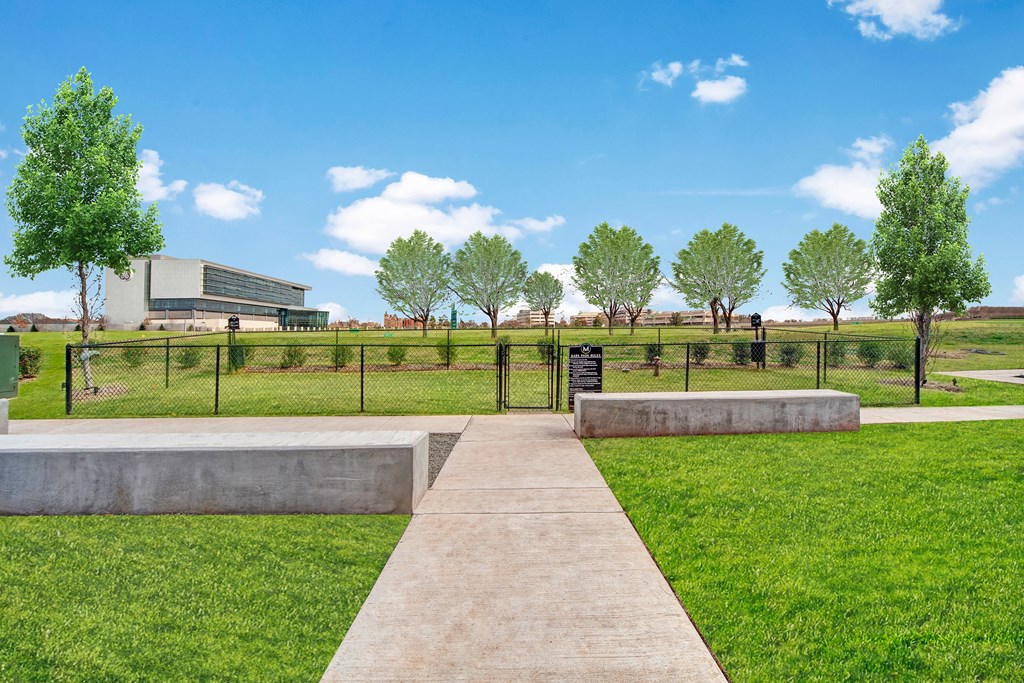 Path leading to a fenced grassy area here at The Metropolitan Apartment Homes with trees and a modern building in the background. Bright blue sky and clouds, conveying a peaceful, spacious feel.