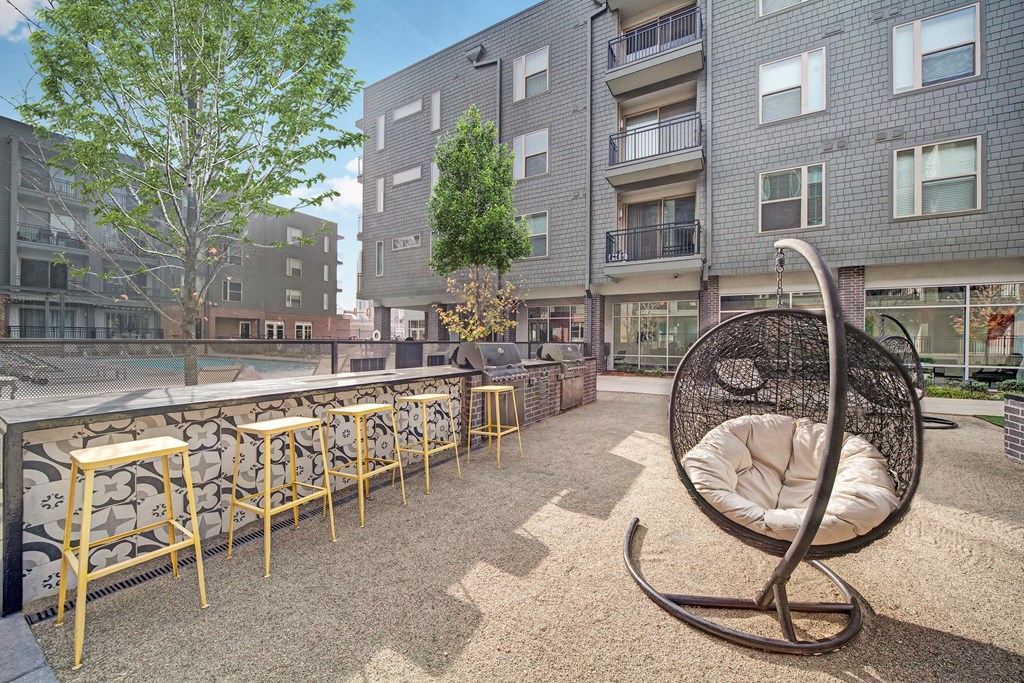 Outdoor patio here at The Metropolitan Apartment Homes with modern design, featuring a hanging wicker chair, high bar stools, and decorative tiled ledge. Surrounded by apartment buildings.