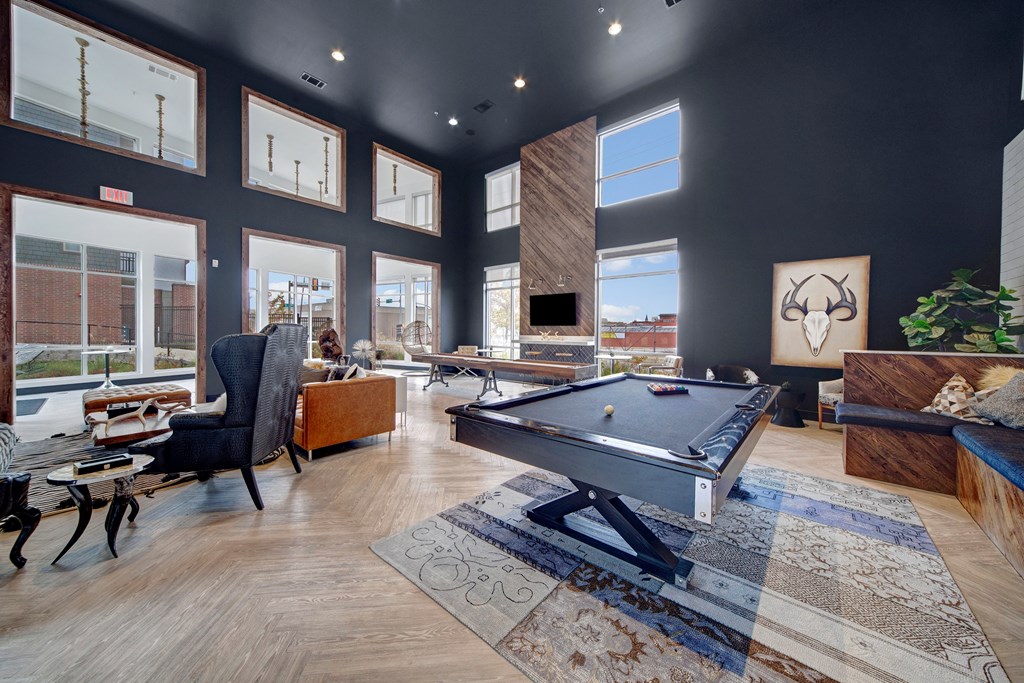 Modern game room here at The Metropolitan Apartment Homes with a sleek pool table in the center, surrounded by stylish seating, a large rug, and wall art. Bright windows create an inviting atmosphere.