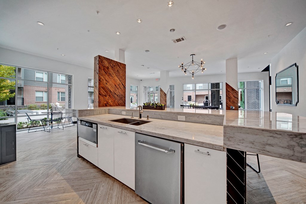 Modern kitchen here at The Metropolitan Apartment Homes with marble countertops, white cabinets, and a double sink. Wooden accents and geometric chandelier create a stylish, open ambiance. Large windows provide natural light and outside view.
