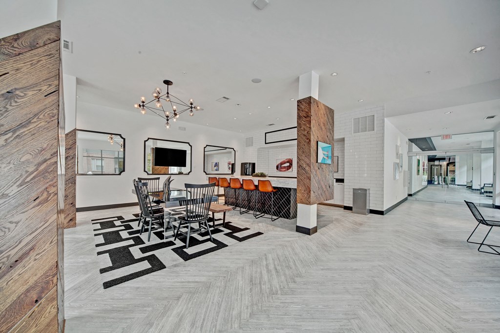 Modern communal space here at The Metropolitan Apartment Homes with a black and white geometric rug, black dining chairs around a glass table, a bar with orange stools, and contemporary lighting.