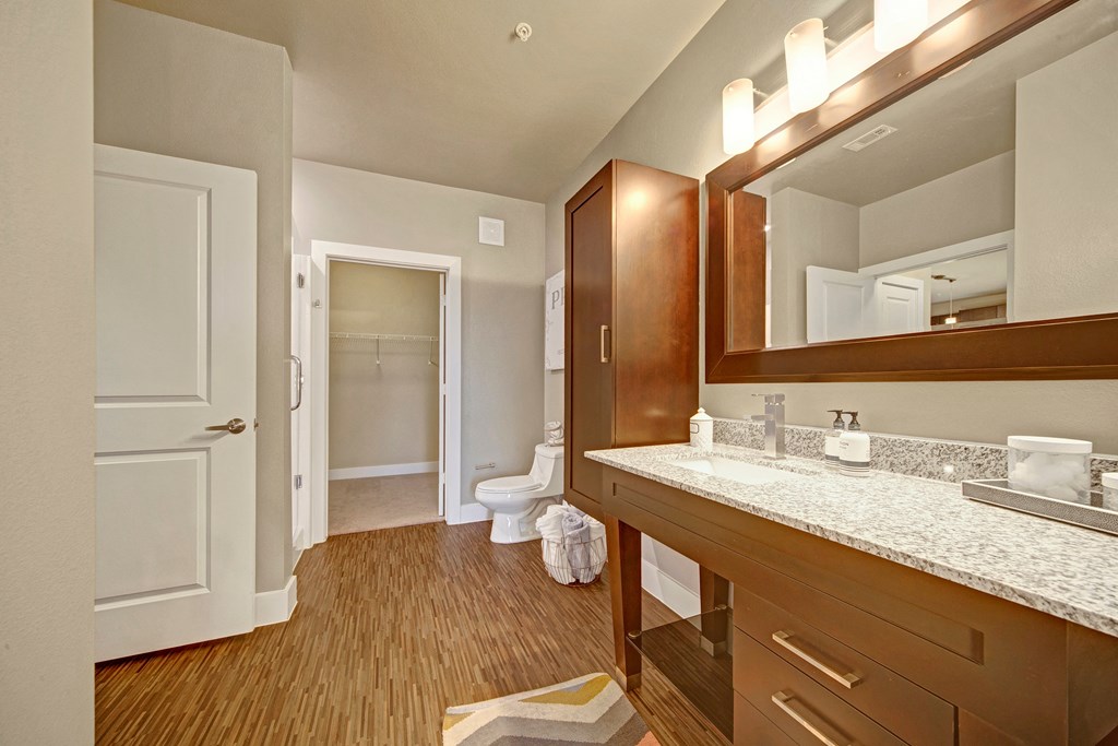 Spacious bathroom here at The Metropolitan Apartment Homes with wood flooring, large granite countertop vanity, and illuminated mirror. Open closet on the left, white towel on towel holder.