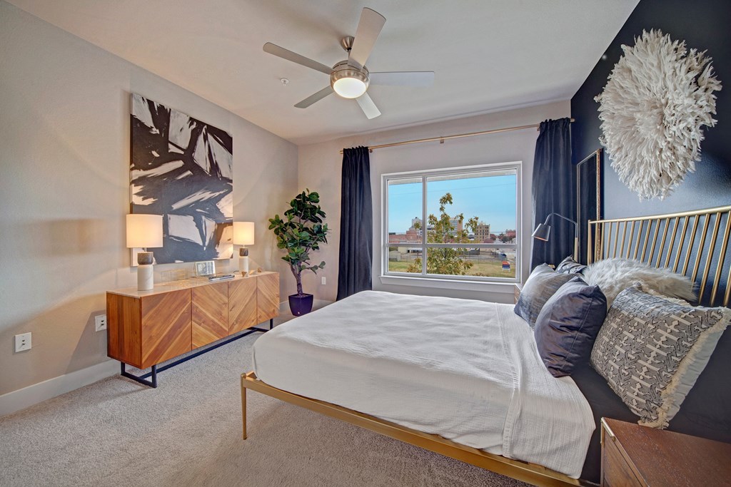 Modern bedroom here at The Metropolitan Apartment Homes with a large window, black accent wall, and abstract art. Features a wooden dresser, cozy bed, fluffy white wall decor, potted plant, and ceiling fan.