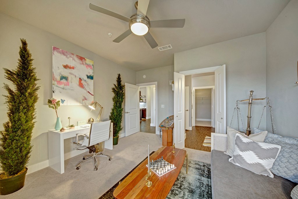 A cozy room here at The Metropolitan Apartment Homes with a modern design features a white desk with a lamp, abstract art, two tall plants, a wooden coffee table with chess, and a gray sofa with pillows.