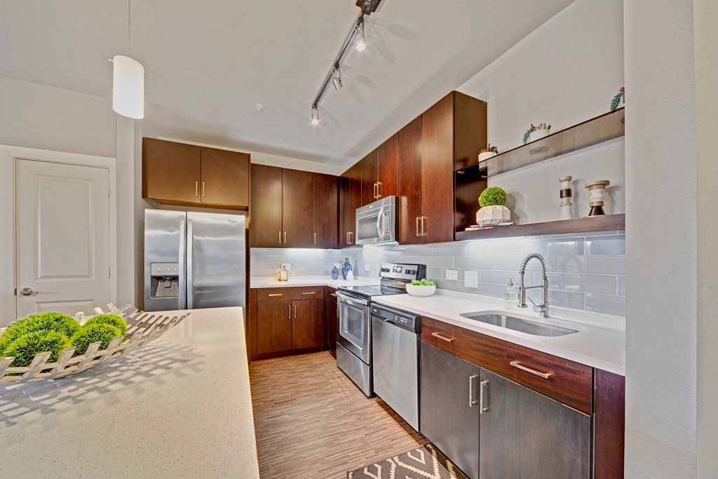 Modern kitchen here at The Metropolitan Apartment Homes with dark wood cabinets, stainless steel appliances, and white countertops. Soft lighting and decorative plants create a cozy atmosphere.