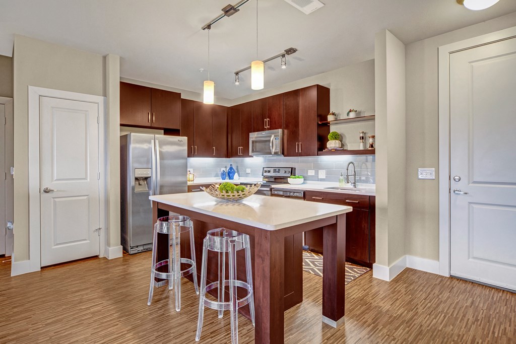 Modern kitchen here at The Metropolitan Apartment Homes with dark wood cabinets, stainless steel appliances, and an island with clear stools. Bright lighting creates a welcoming atmosphere.