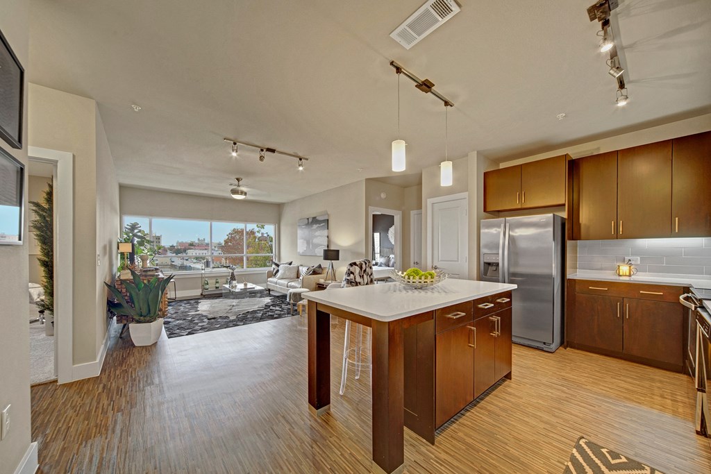 Modern open-plan living area here at The Metropolitan Apartment Homes with wooden floors, featuring a kitchen island, dark cabinets, cozy living room with large windows, and stylish decor.