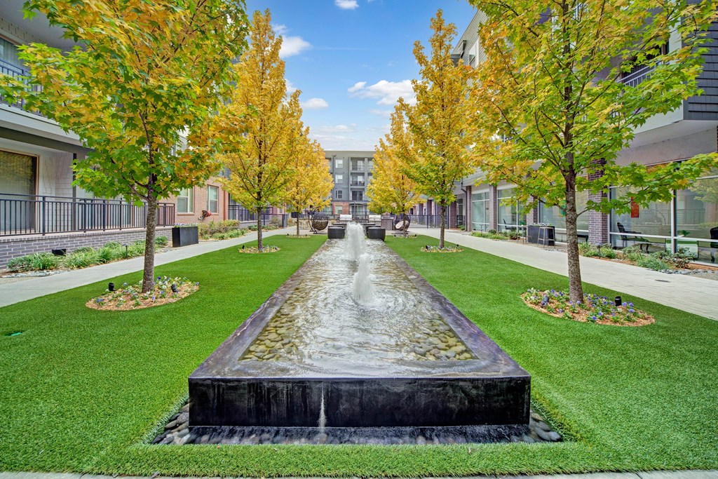 A serene courtyard here at The Metropolitan Apartment Homes features a central rectangular fountain flanked by rows of lush trees with fall foliage. Manicured grass and flower beds line the path.