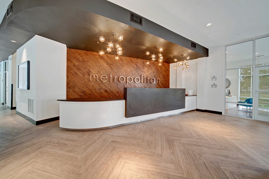 Modern reception area here at The Metropolitan Apartment Homes with a sleek, curved desk against a rustic wood wall featuring "metropolitan" in silver. Chic lighting and a calm, inviting atmosphere.