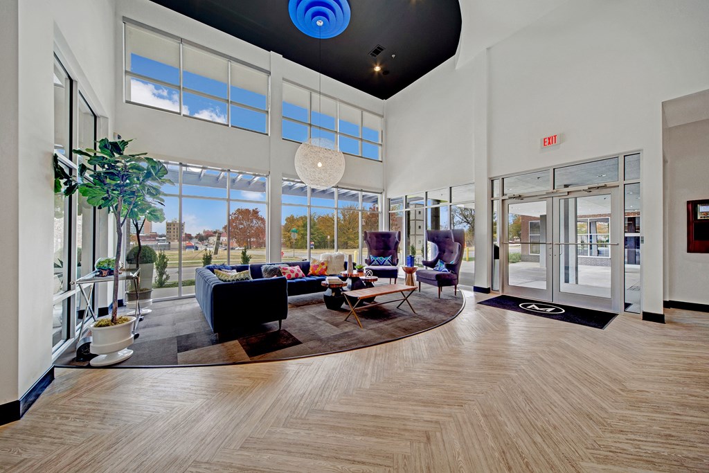 Modern The Metropolitan Apartment Homes lobby with large windows and ample natural light. Features cozy seating, a round coffee table, potted plants, and a stylish blue ceiling.