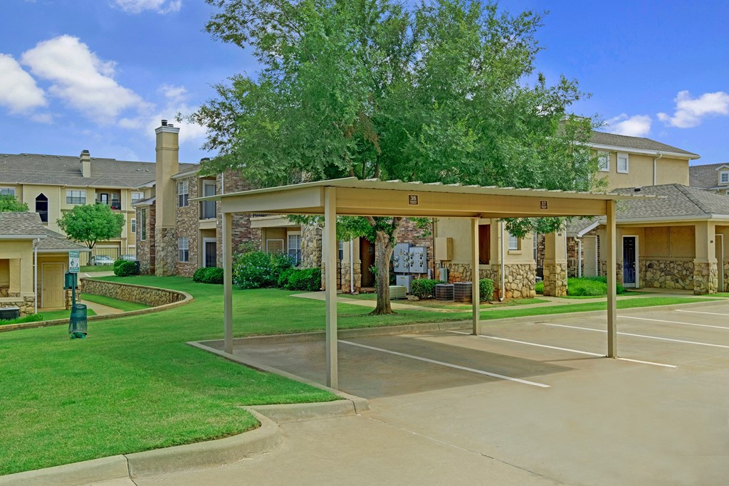 Suburban Quail Landing Apartment Homes with modern architecture, stone facades, and green lawns. Covered parking in foreground, tree casting shade. Bright, sunny day.