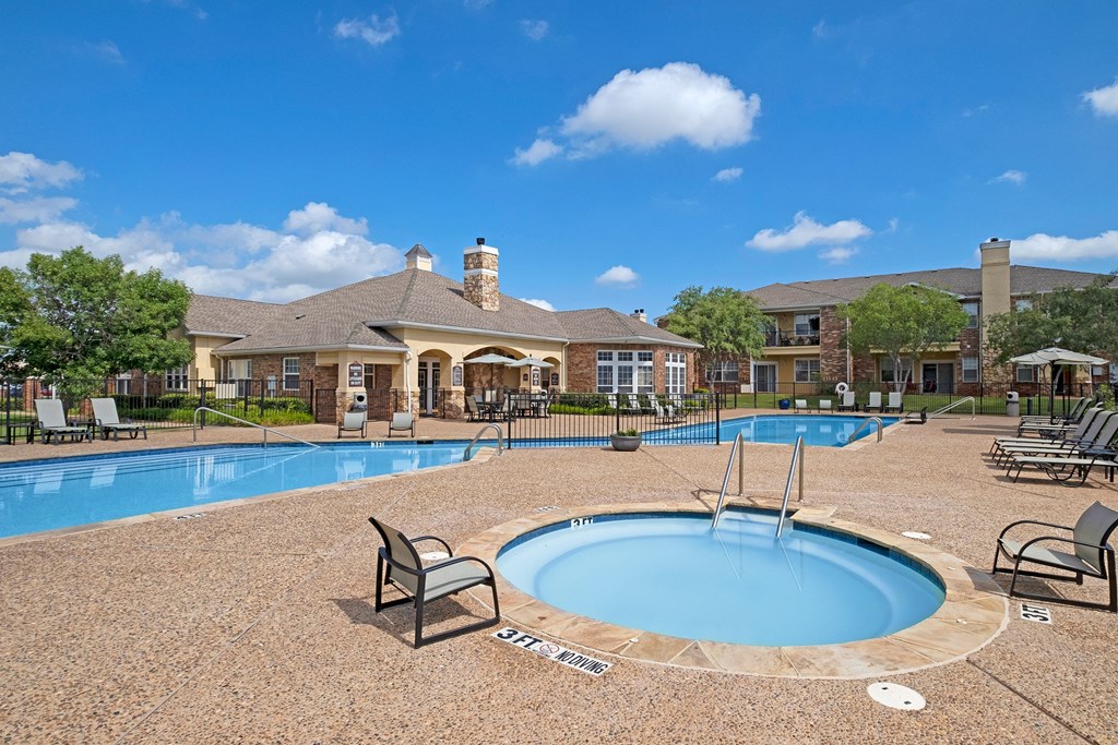 Sunny pool area here at Quail Landing Apartment Homes with a rectangular swimming pool and circular hot tub, surrounded by lounge chairs. Apartments and green trees in the background. Relaxing ambiance.