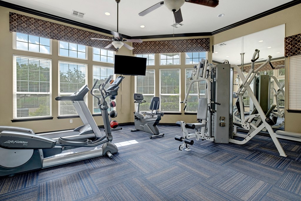 A bright gym room here at Quail Landing Apartment Homes with cardio machines, a multi-gym, and a TV. Large windows let in natural light, creating an inviting, energetic atmosphere.