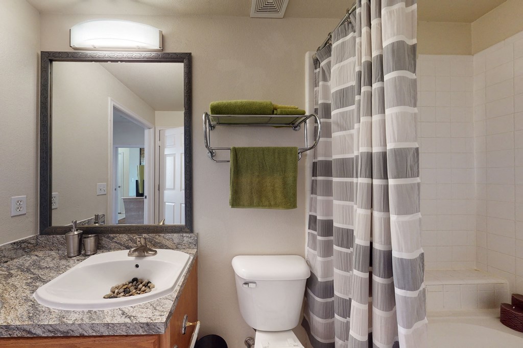 Modern bathroom here at Quail Landing Apartment Homes with granite countertop, sink adorned with pebbles, green towels on a metal rack, and a striped shower curtain. Calm, clean ambiance.