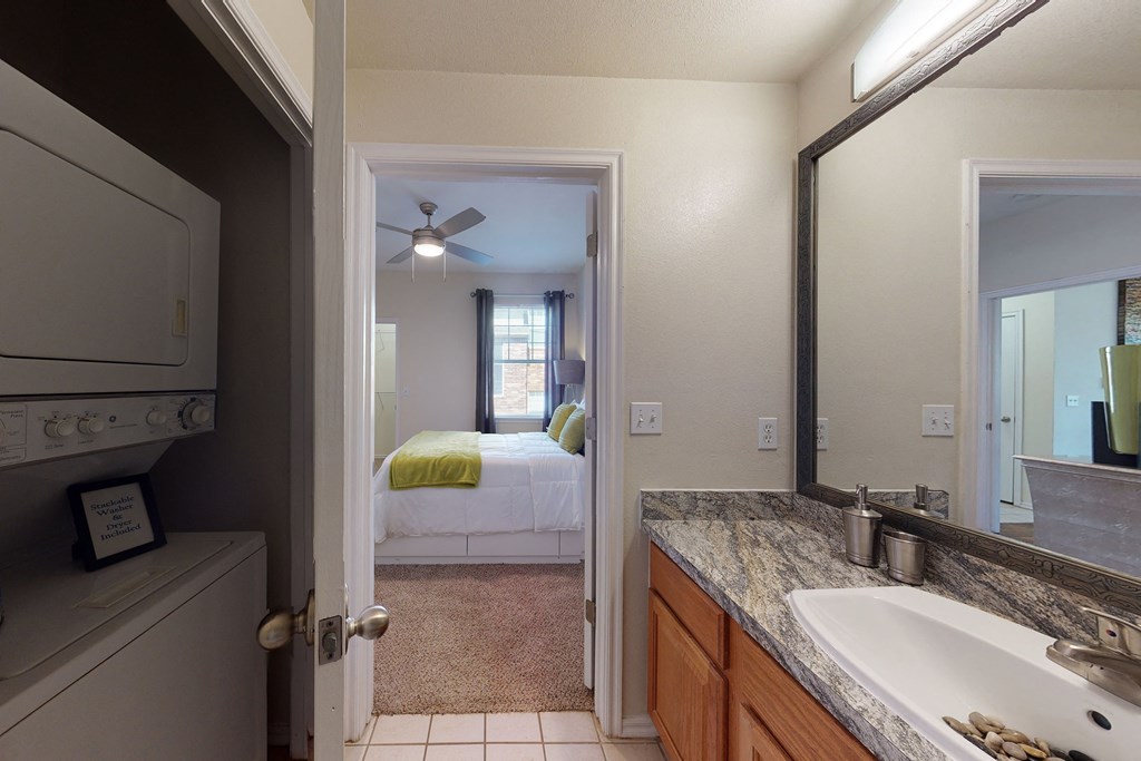 View from a bathroom here at Quail Landing Apartment Homes with a granite countertop and sink, looking into a bright bedroom with white bedding and green accents. A stacked washer-dryer unit is to the left.