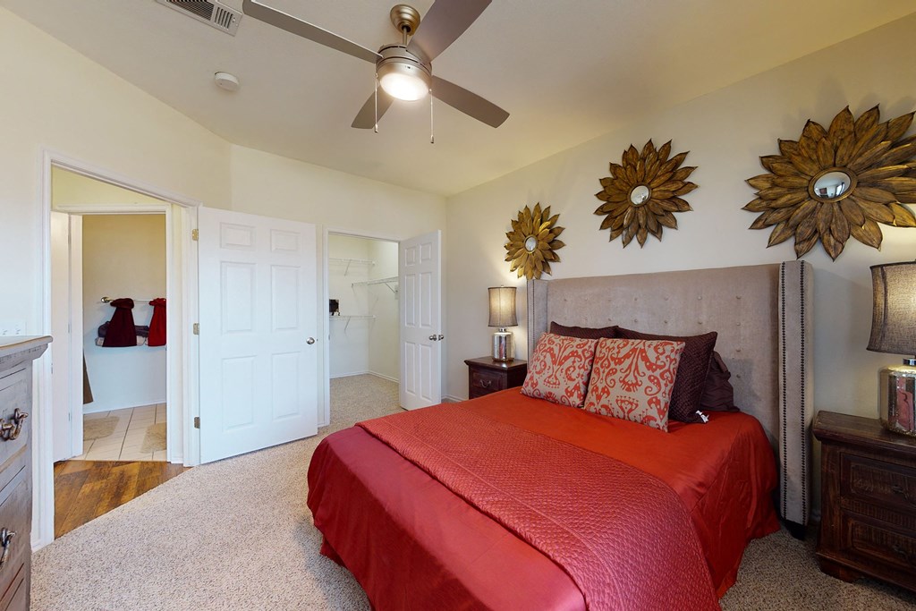 Bedroom here at Quail Landing Apartment Homes with red bedding and two floral metal wall art pieces above the bed. A ceiling fan is centered, with open doors leading to a bathroom and closet.