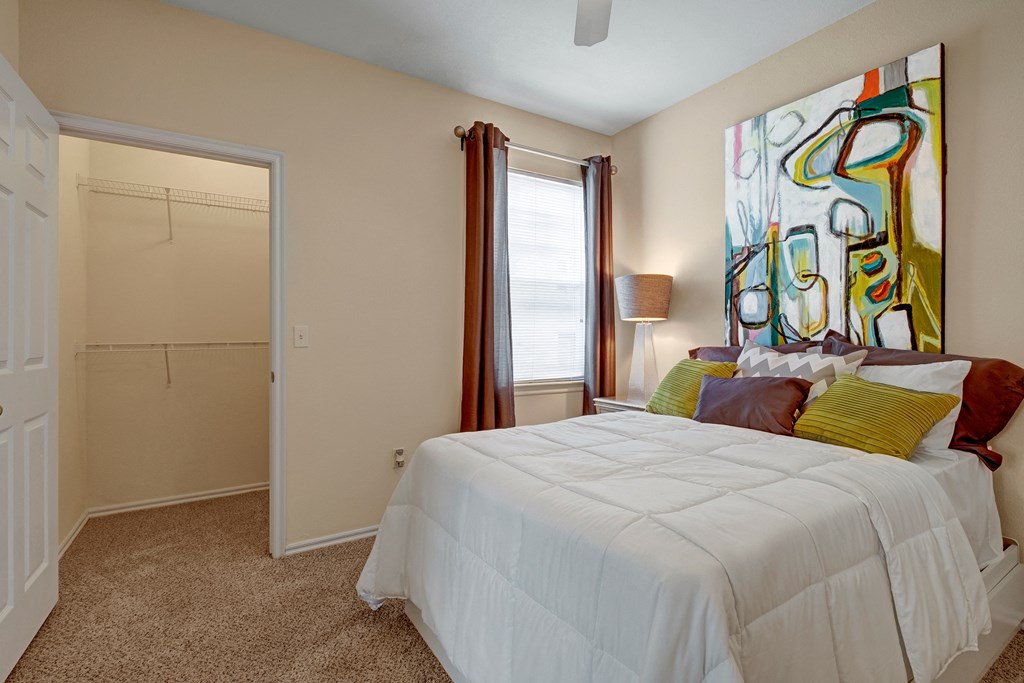 A cozy bedroom here at Quail Landing Apartment Homes with a large abstract painting above the bed, featuring colorful shapes. The bed has white bedding, with green and brown pillows. There's soft lighting from a lamp beside the bed, and a window with brown curtains. An open closet door is visible, revealing an empty storage space. The tone is warm and inviting.