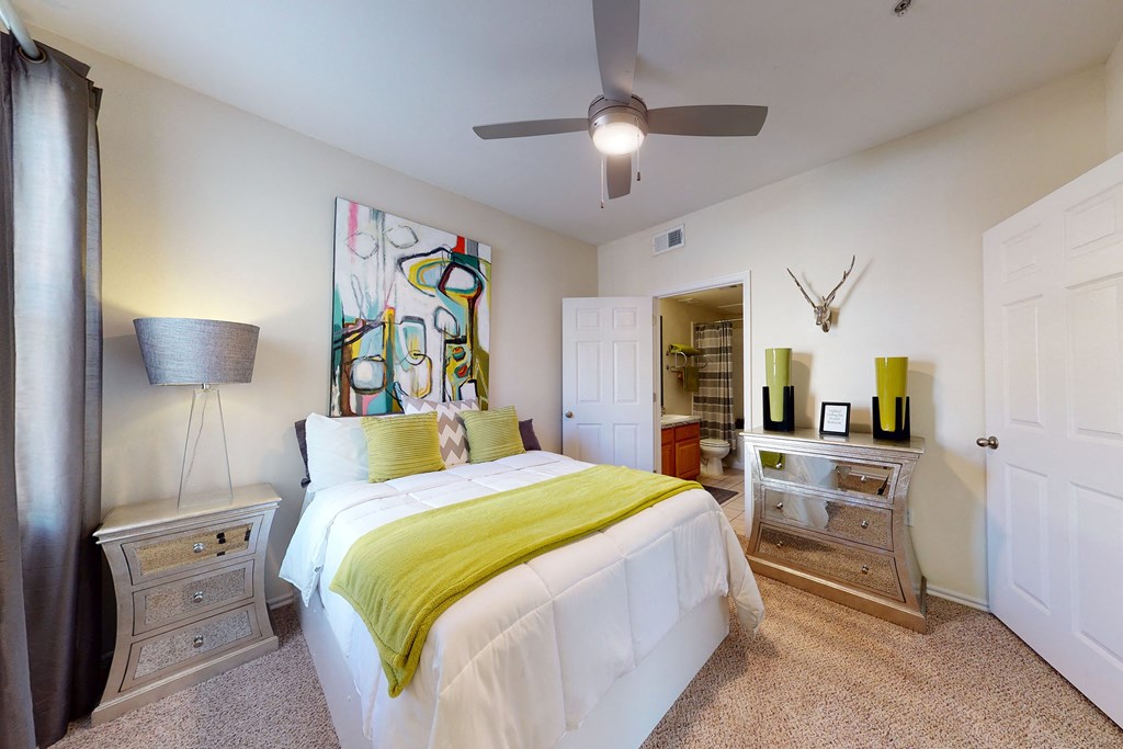 Cozy bedroom here at Quail Landing Apartment Homes with a bed featuring a green throw blanket and abstract art above. Mirrored nightstands, beige carpet, and a ceiling fan enhance the space.