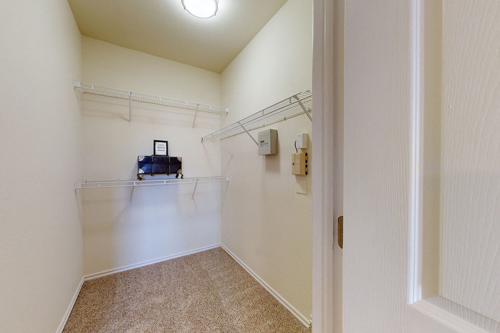 Walk-in closet here at Quail Landing Apartment Homes with carpeted floor, beige walls, and white wire shelving. A small framed picture and box sit on the middle shelf, under a ceiling light.
